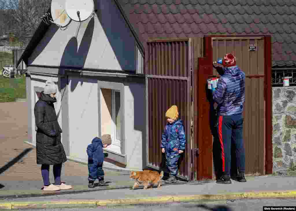 Daria (majtas) është fotografuar më 30 mars 2023, duke shikuar burrin e saj Maxim që po lyente portën e shtëpisë së tyre, teksa afër tyre fëmijët e tyre lozin. Një vit më parë, zrupat e pajetë të cilëve ishin gjetur edhe në rrugën pranë shtëpisë së tyre.