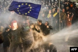 Imagine definitorie pentru protestele din 2023, împotriva legii agenților străini: Poliția încearcă să doboare cu tunuri cu apă o femeie care ține un steag al Uniunii Europene. Tbilisi, martie 2023.