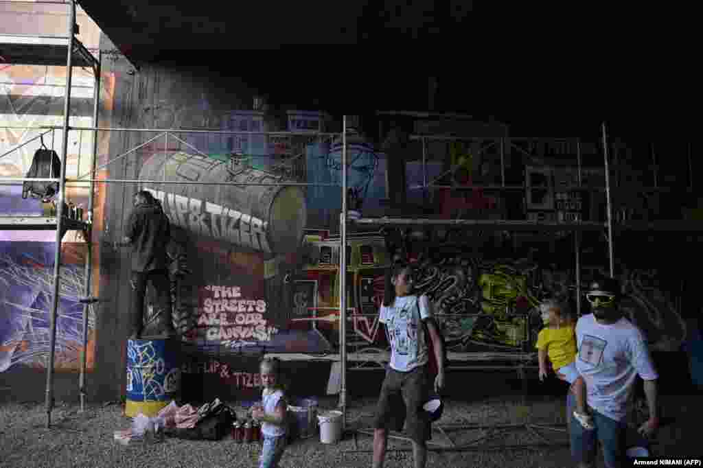 Vizitatori trecând pe lângă artistul din Insulele Canare fEOfL!P în timp ce acesta lucrează la creația sa. Organizatorii scot în evidență puterea artei stradale ca instrument de educație și transformare socială.