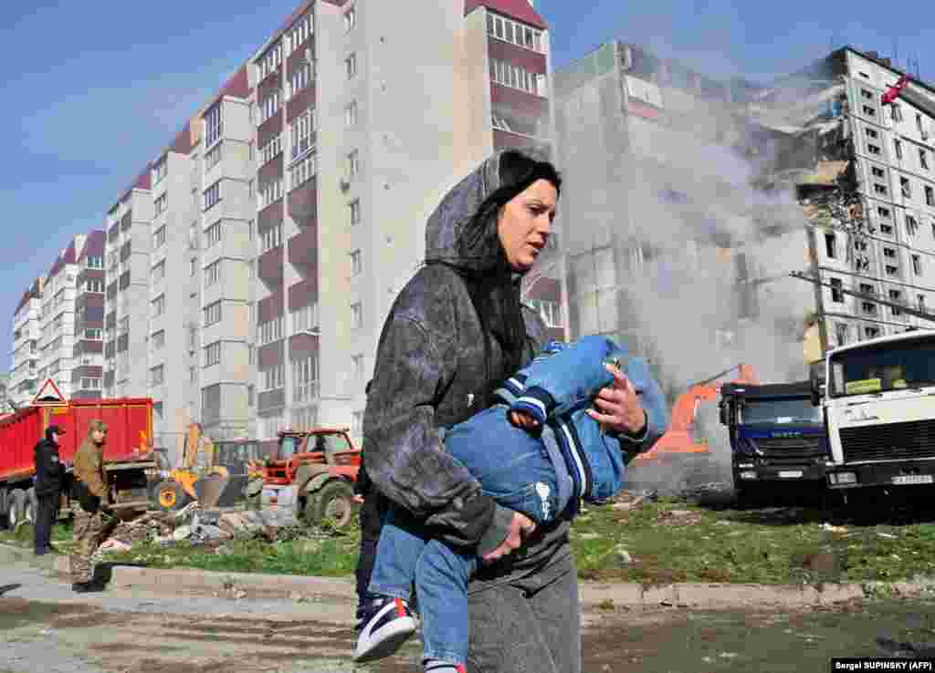 A woman carries a child past the smoldering residential building in Uman after it was struck by Russian missiles.