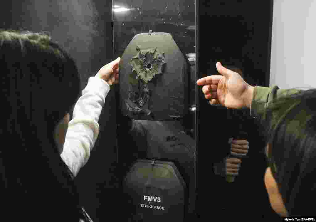 Vizitatorii expoziției examinează urma unui proiectil pe o vestă anti-glonț. Site-ul web al expoziției arată &bdquo;veste antiglonț care au salvat viața cuiva pe câmpul de luptă, care au o a doua șansă de a salva o viața cuiva cu ajutorul unor artiști celebri&rdquo;.