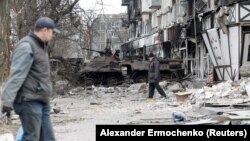 Locuitorii merg în apropierea unui vehicul blindat ars și a unui bloc de apartamente distrus în timpul conflictului dintre Ucraina și Rusia, în orașul-port din sudul asediat Mariupol, Ucraina, 25 martie 2022.