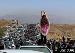 Жена без забрадка стои върху автомобил на фона на хиляди хора, които си проправят път към гробището в Сакез, родния град на Махса Амини в западната иранска провинция Кюрдистан, 26 октомври 2022 г.
