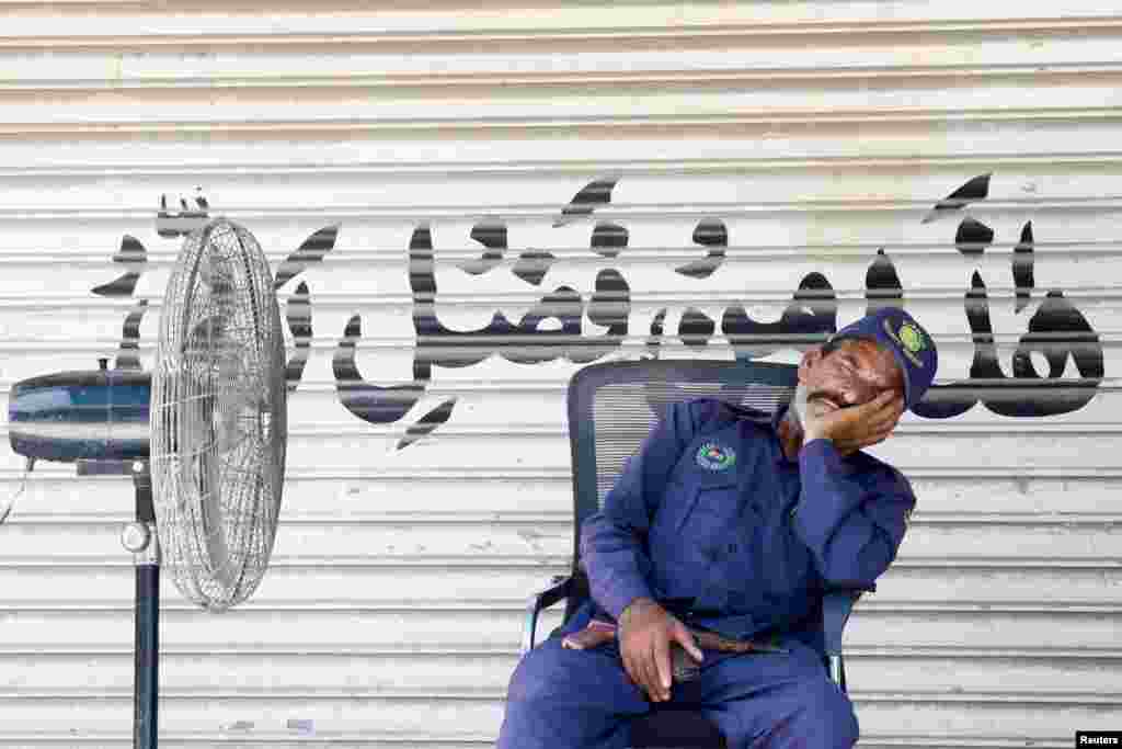 Охранник дремлет рядом с напольным вентилятором перед закрытым магазином, на роллете которого надпись "Это — по милости моего Господа". Карачи, Пакистан, 22 апреля 2025 года. REUTERS/Akhtar Soomro