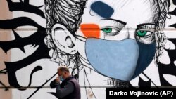 Një burrë rregullon maskën mbrojtëse teska kalon pranë një murali me një portret po ashtu me maskë mbrojtëse, Mitrovicë.
