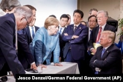 O imagine ca aceasta valorează chiar mai mult de o mie de cuvinte. Angela Merkel discută cu Donald Trump la summitul G7 din 2018. Președintele francez Emmanuel Macron ține frontul împreună cu Merkel, în timp ce premierul japonez Shinzo Abe privește exasperat.