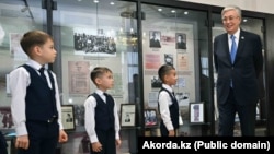 Қасым-Жомарт Тоқаев Үштөбе қаласындағы Кемел Тоқаев атындағы мектепте оқушылармен бірге тұр. Арғы жағында әйнектің астында – презиенттің әкесі, майдангер жазушы жайлы деректер мен фотолар ілінген. Жетісу облысы, 27 қазан, 2023 жыл.