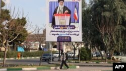 ქალი საარჩევნო ბილბორდის წინ, რომელზეც გამოსახულია უზენაესი ლიდერი, აიათოლა ალი ხამენეი. ყომი, 22 თებერვალი
